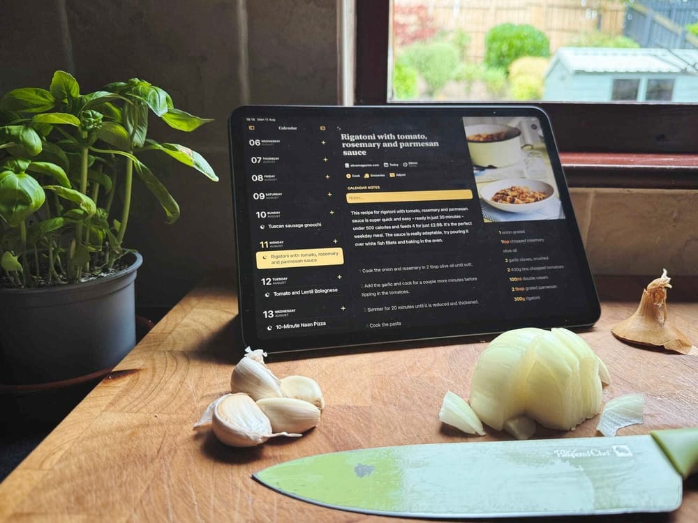 A kitchen countertop displays a chopping board with garlic, a half-sliced onion, a knife, and a digital tablet showing a recipe, with a potted basil plant in the background.