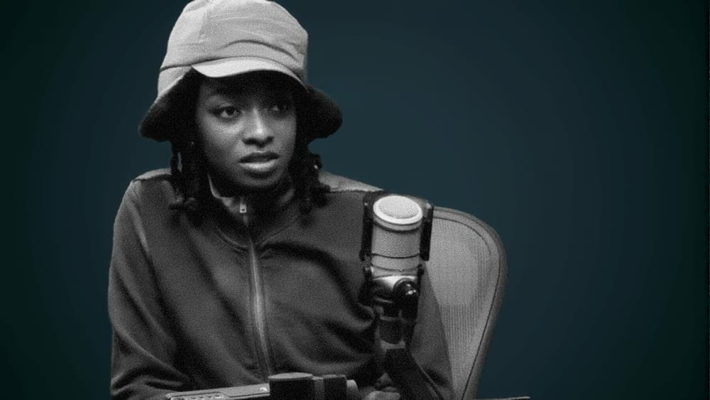 Little Simz wearing a bucket hat sits in front of a microphone in a recording studio.