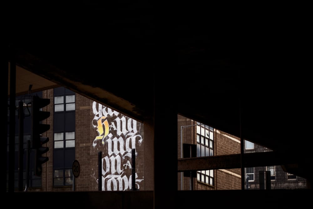 White blackletter lettering painted on a brick building, glimpsed through gaps in a dark car park structure, with traffic lights in the left foreground.