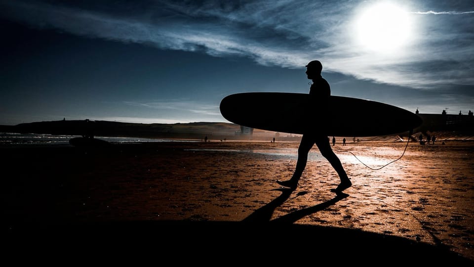 Silhouette of a surfer carrying a longboard across a wet beach, backlit by a low bright sun.